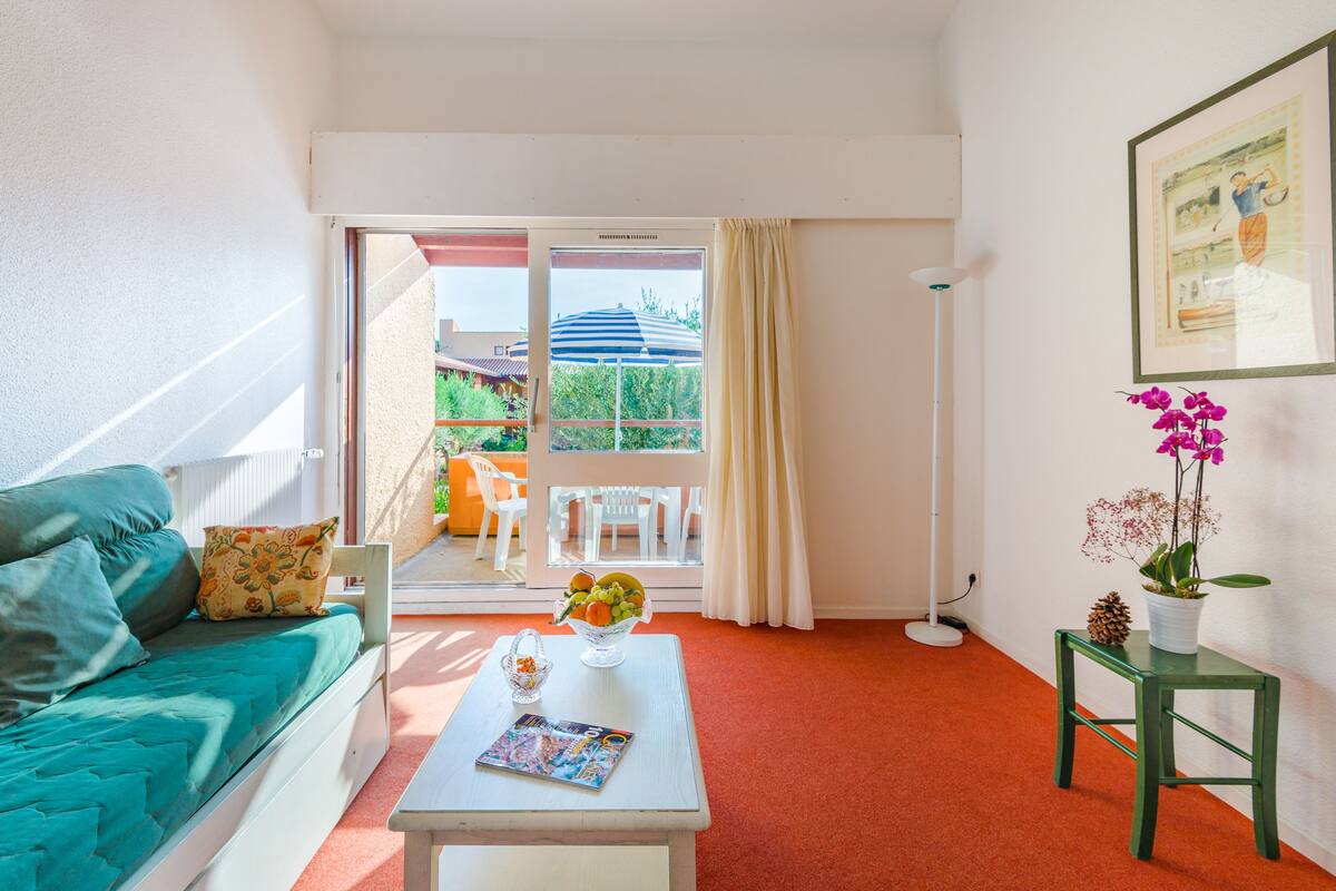 Ein helles Zimmer mit einem türkisfarbenen Sofa, einem weißen Couchtisch mit einer Zeitschrift, einem kleinen Tisch mit einem Blumentopf und einem großen Fenster mit Blick auf Gartenmöbel.