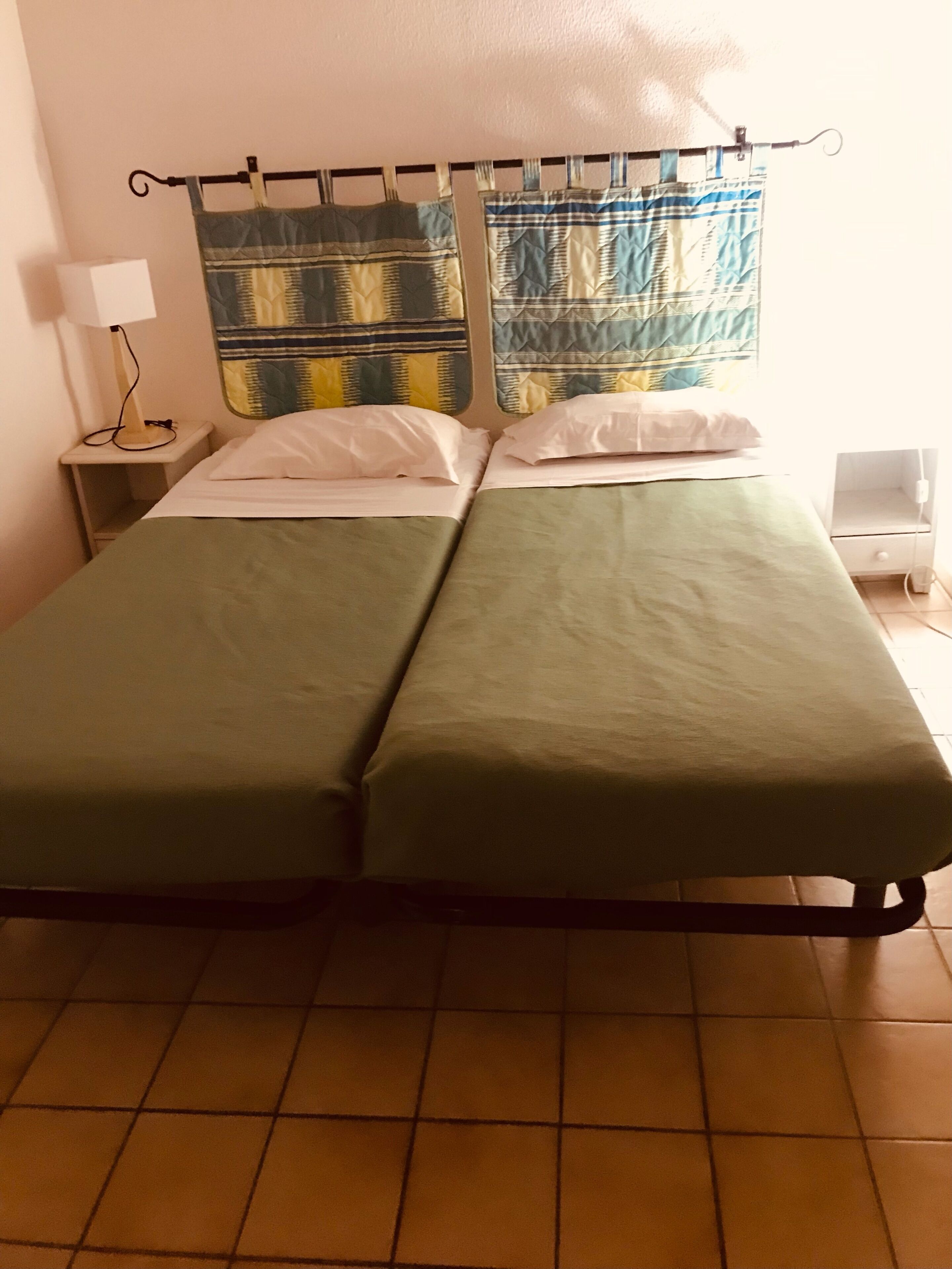 Ein Doppelbett mit grüner Bettwäsche, ein Nachttisch mit Lampe und ein Fenster mit gemusterten Vorhängen.