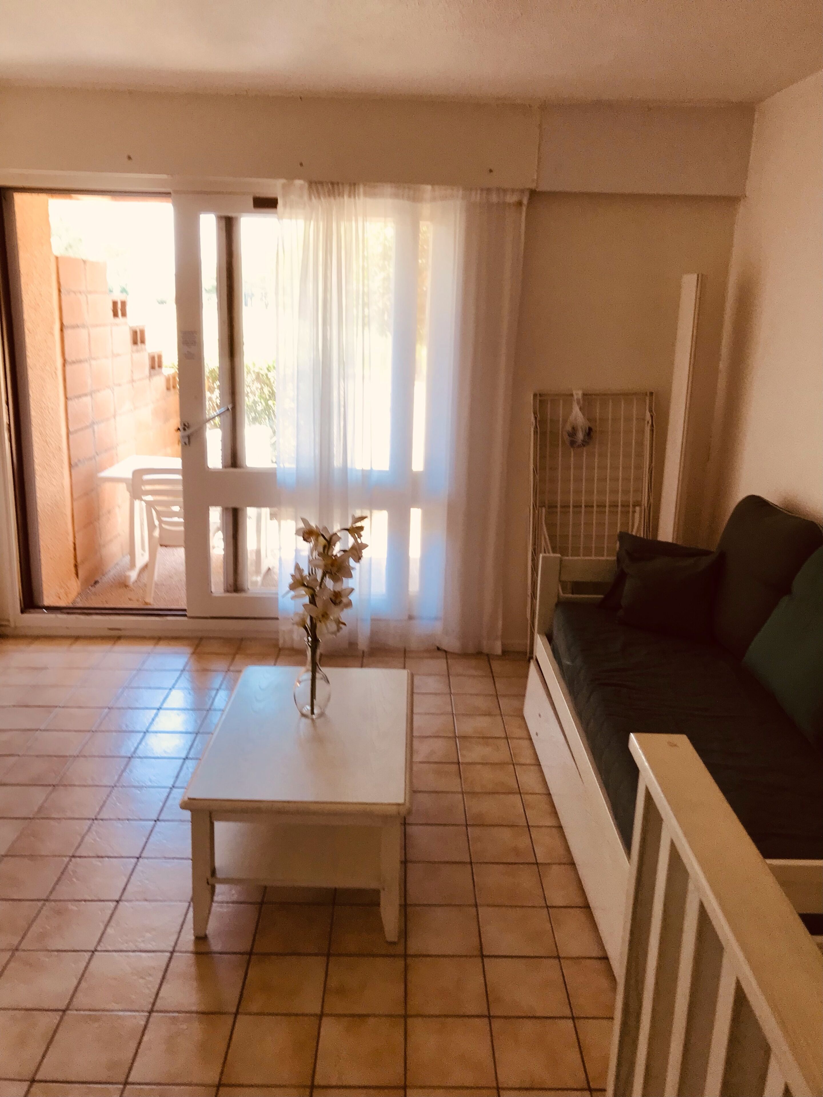 Une pièce avec un sol carrelé, une table basse blanche ornée d’un vase, un canapé foncé et une porte-fenêtre coulissante donnant sur l’extérieur.