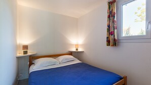 A single bed with a blue bedspread, two bedside lamps, a window with floral curtains, and a white wall.