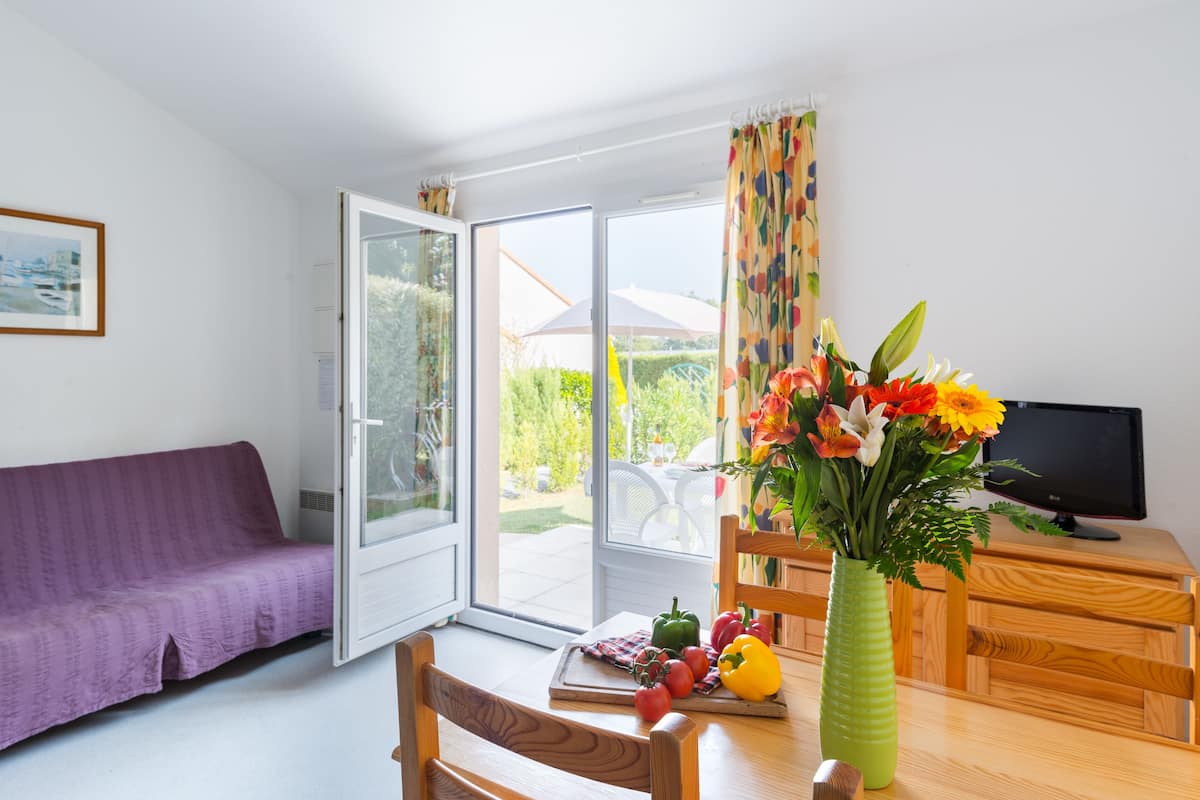 Ein Zimmer mit einem violetten Sofa, einem Esstisch mit Blumen in einer Vase, einem Fernseher und einer Glastür, die in einen Garten führt.