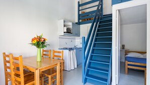 A compact room with a wooden dining table, chairs, a small kitchen area, and a blue staircase leading to a loft bed.