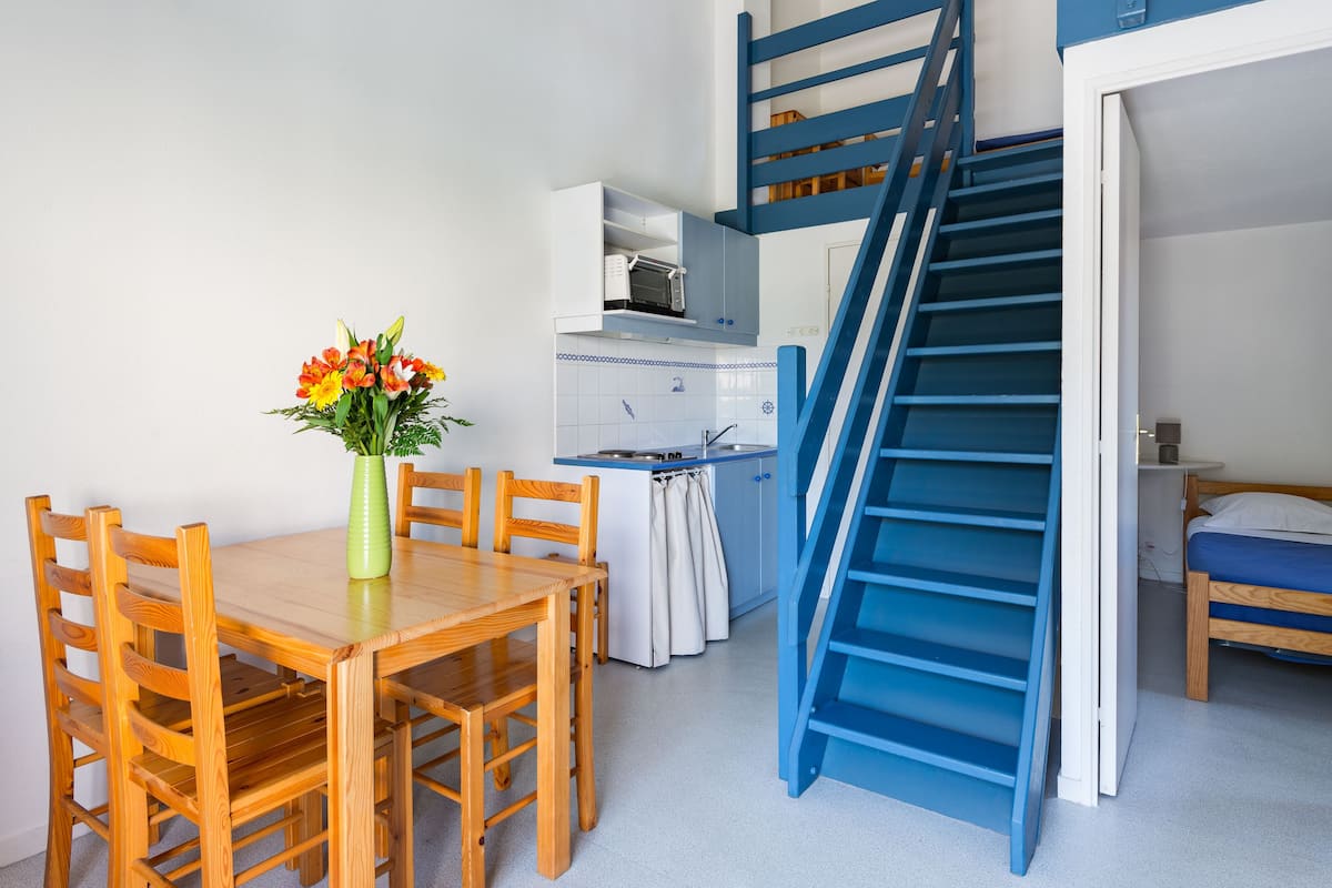 Ein gemütlicher Raum mit Esstisch aus Holz, Stühlen, einer kleinen Küchenzeile und einer blauen Treppe, die zu einem Schlafplatz im Podest führt.