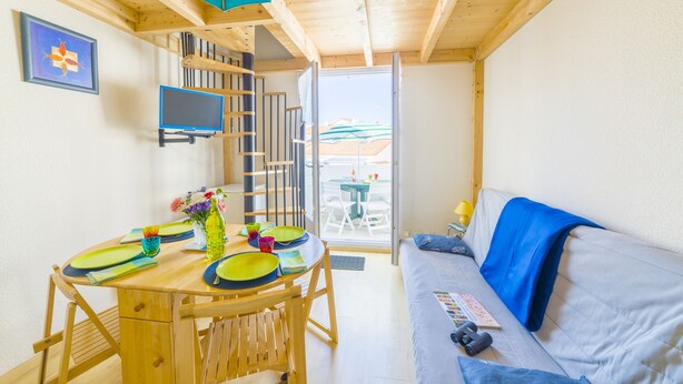 A cozy room with a wooden ceiling, a dining table with colorful plates, a sofa with a blue blanket, and a window with a view of a building. - Résidence Lagrange Vacances Les Fermettes de la Mer (Saint-Jean-de-Monts)