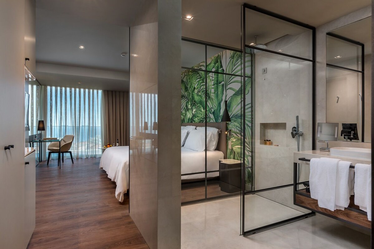 Une chambre d’hôtel moderne dotée d’un balcon, d’un lit, d’un bureau et d’une salle de bain avec un grand miroir et une douche.