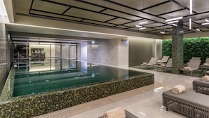 Un espace piscine intérieur moderne, doté d’une paroi vitrée, de chaises longues et d’un comptoir d’accueil.