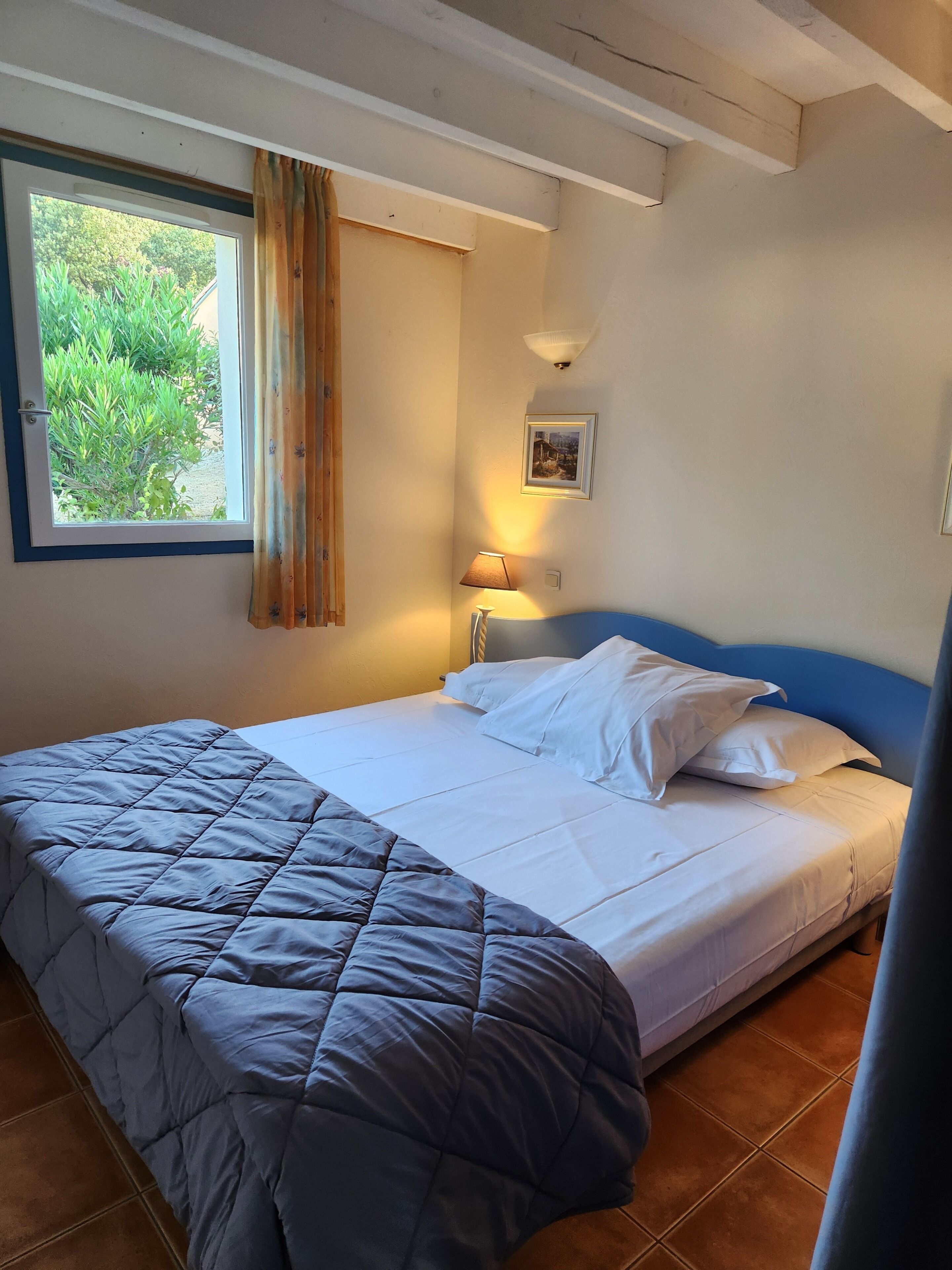 A single bed with a blue headboard, a window with curtains, a wall-mounted lamp, and a framed picture.