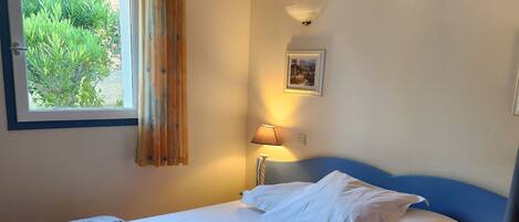 A single bed with a blue headboard, a window with curtains, a wall-mounted lamp, and a framed picture.