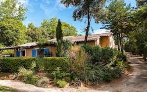 A rustic house with a tiled roof, surrounded by dense greenery and a gravel path.