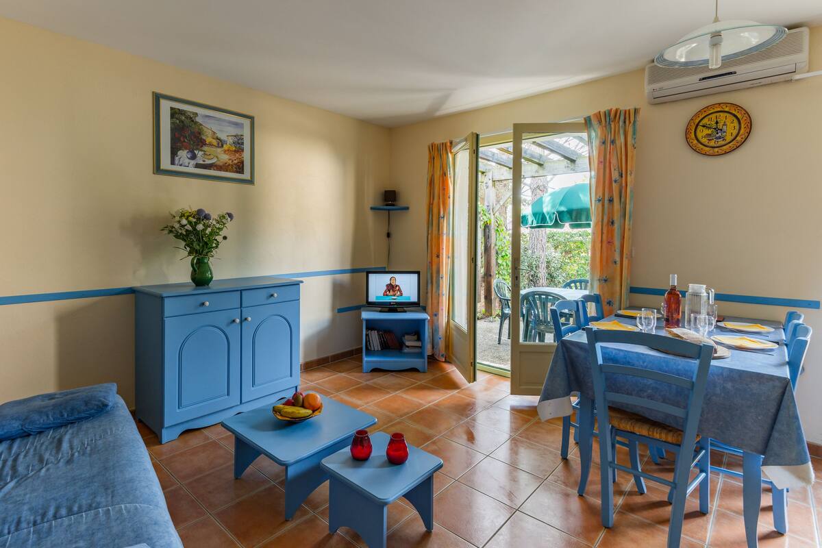 Una sala de estar con un sofá, un televisor, una mesita con fruta y un comedor con sillas y una mesa.