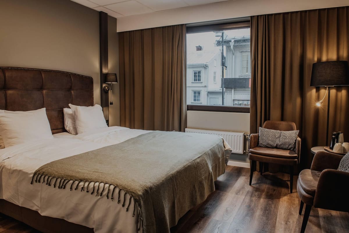 A hotel room with a large bed, a chair, a lamp, and a view of buildings outside the window.