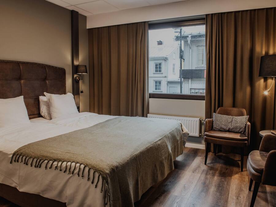 Una habitación de hotel con una cama grande, una silla, una lámpara y vistas a edificios desde la ventana.