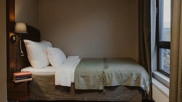 A corner bedroom with a bed, a nightstand, a lamp, a window with curtains, and a radiator.