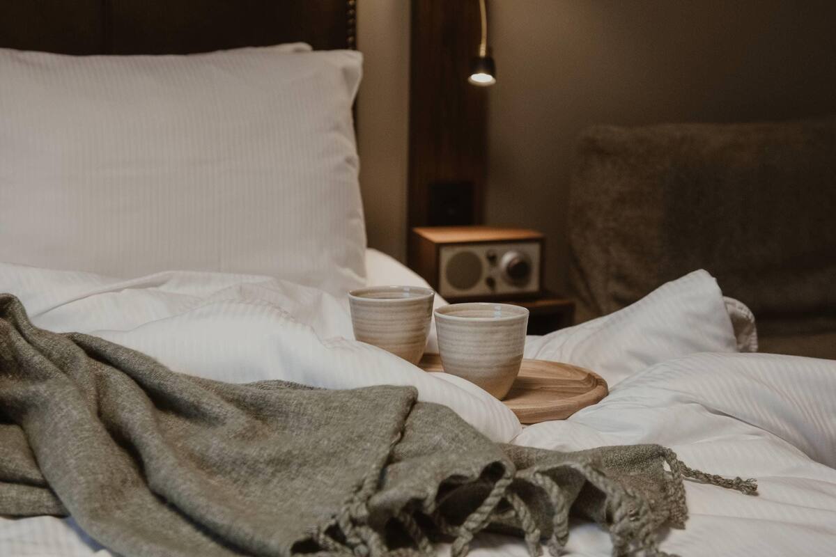 A bed with white bedding, a grey blanket, and two ceramic cups on a wooden tray.