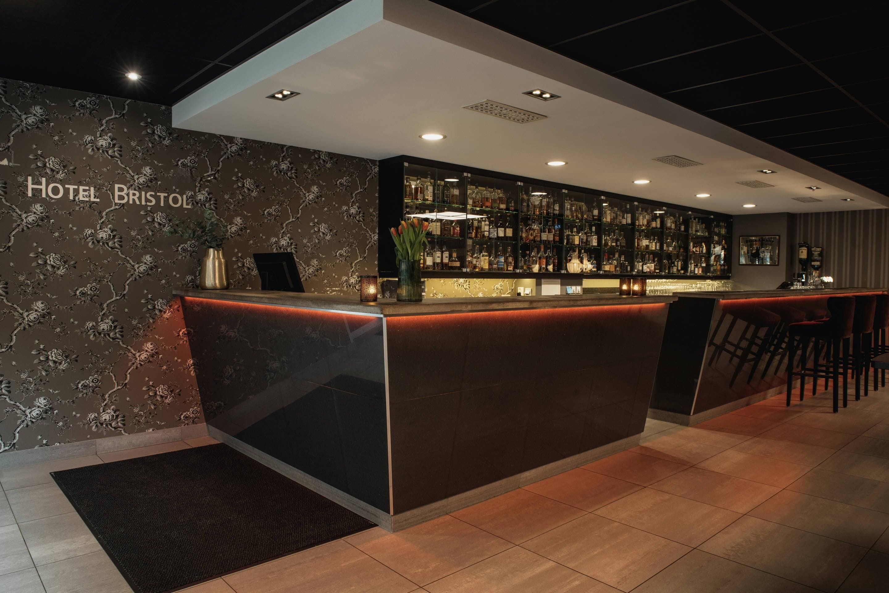 Een bar met een naambordje waarop 'Hotel Bristol' staat, een goed gevulde drankkast en een zithoek met barstoelen.