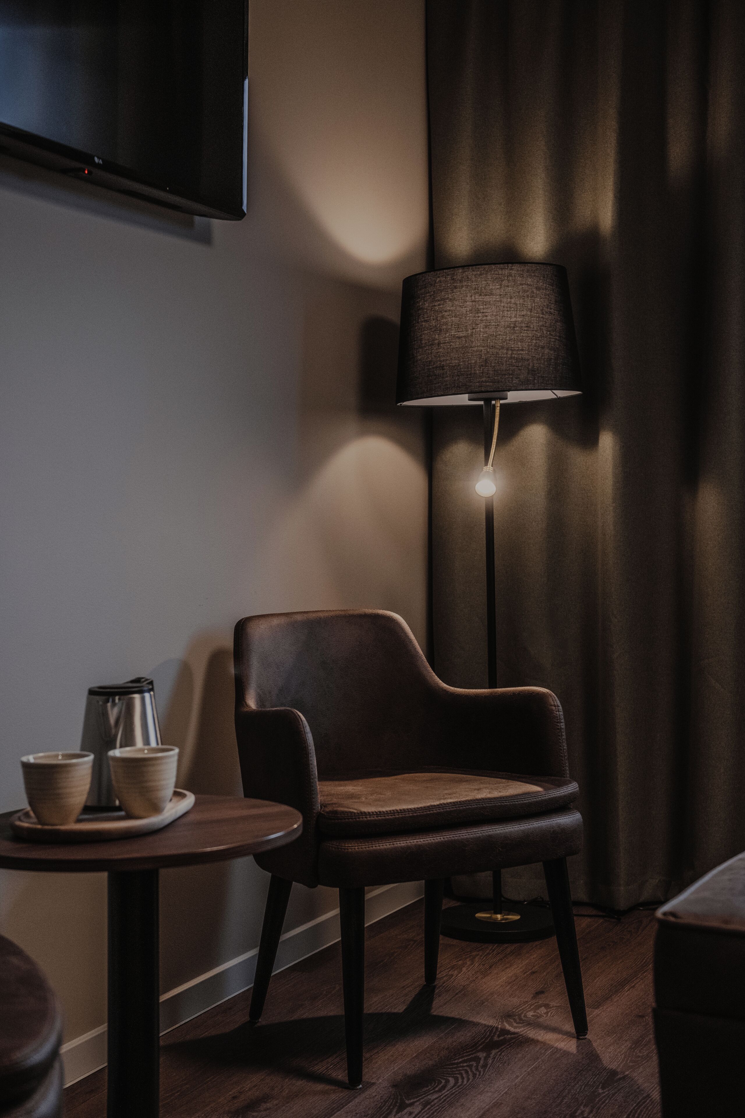 A modern room with a flat-screen TV, a dark-colored armchair, a small round table with a coffee pot and cups, and a floor lamp.