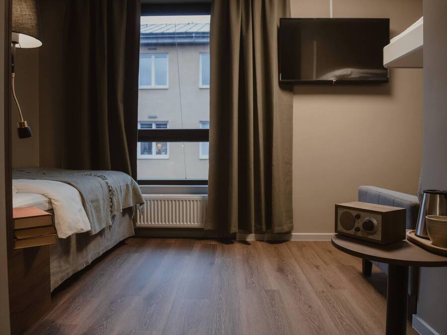 A hotel room with a bed, a table with a radio, a TV, and a window with curtains.