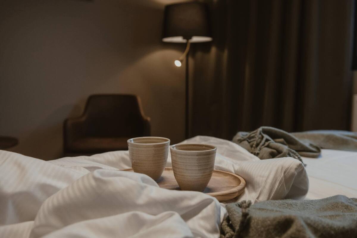 A bed with white bedding, two ceramic cups on a wooden tray, a lamp, and a chair in a dimly lit room.