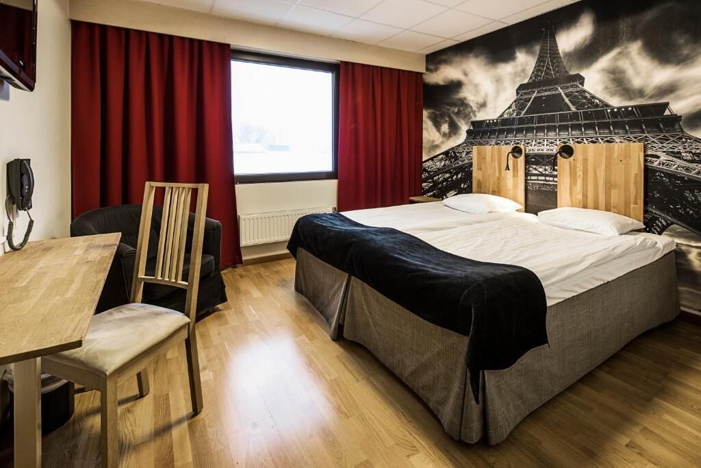 Een hotelkamer met een bed, een bureau, een stoel, een raam met rode gordijnen en een muurschildering van de Eiffeltoren.