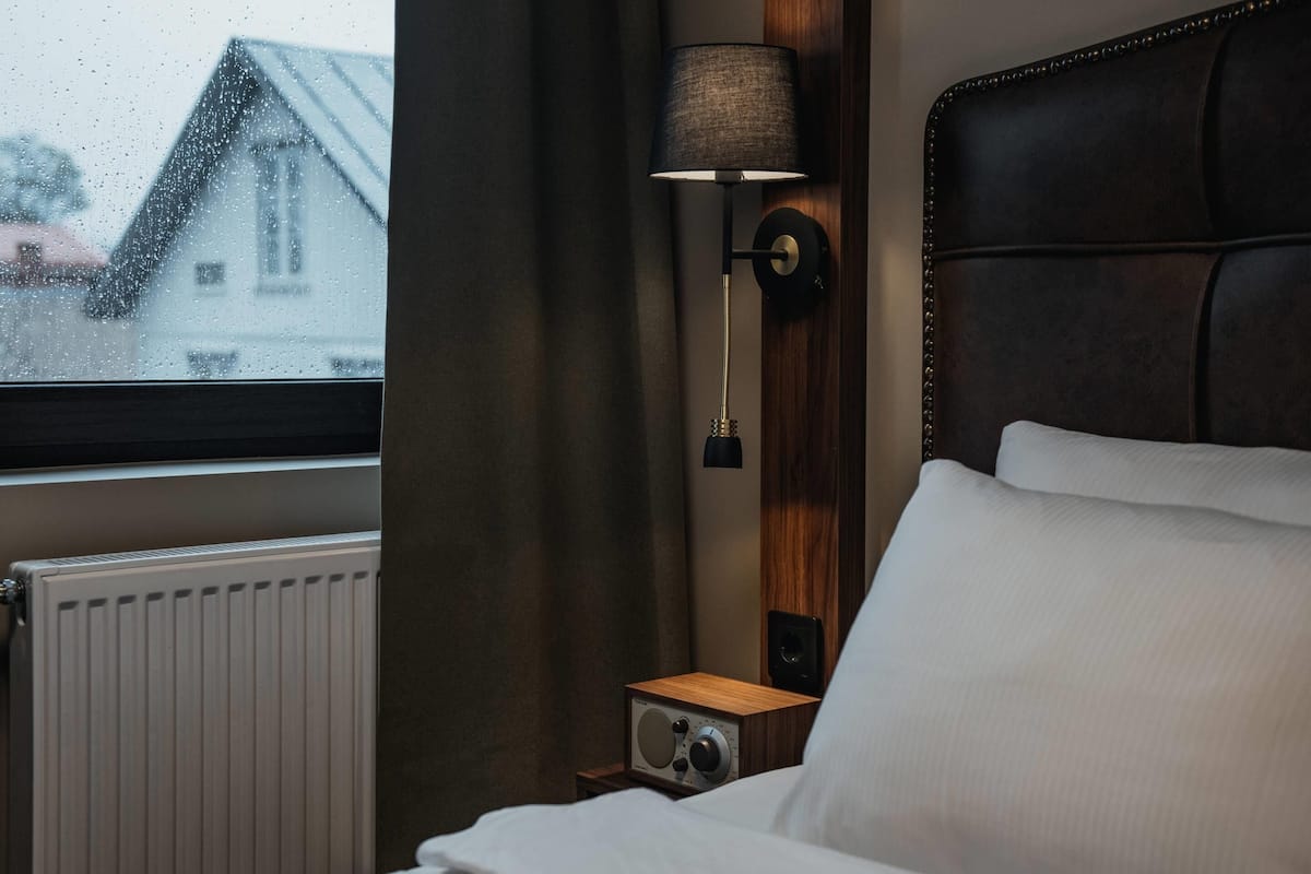 A hotel room with a bed, a nightstand with a radio, a lamp, and a view of a house through the window.