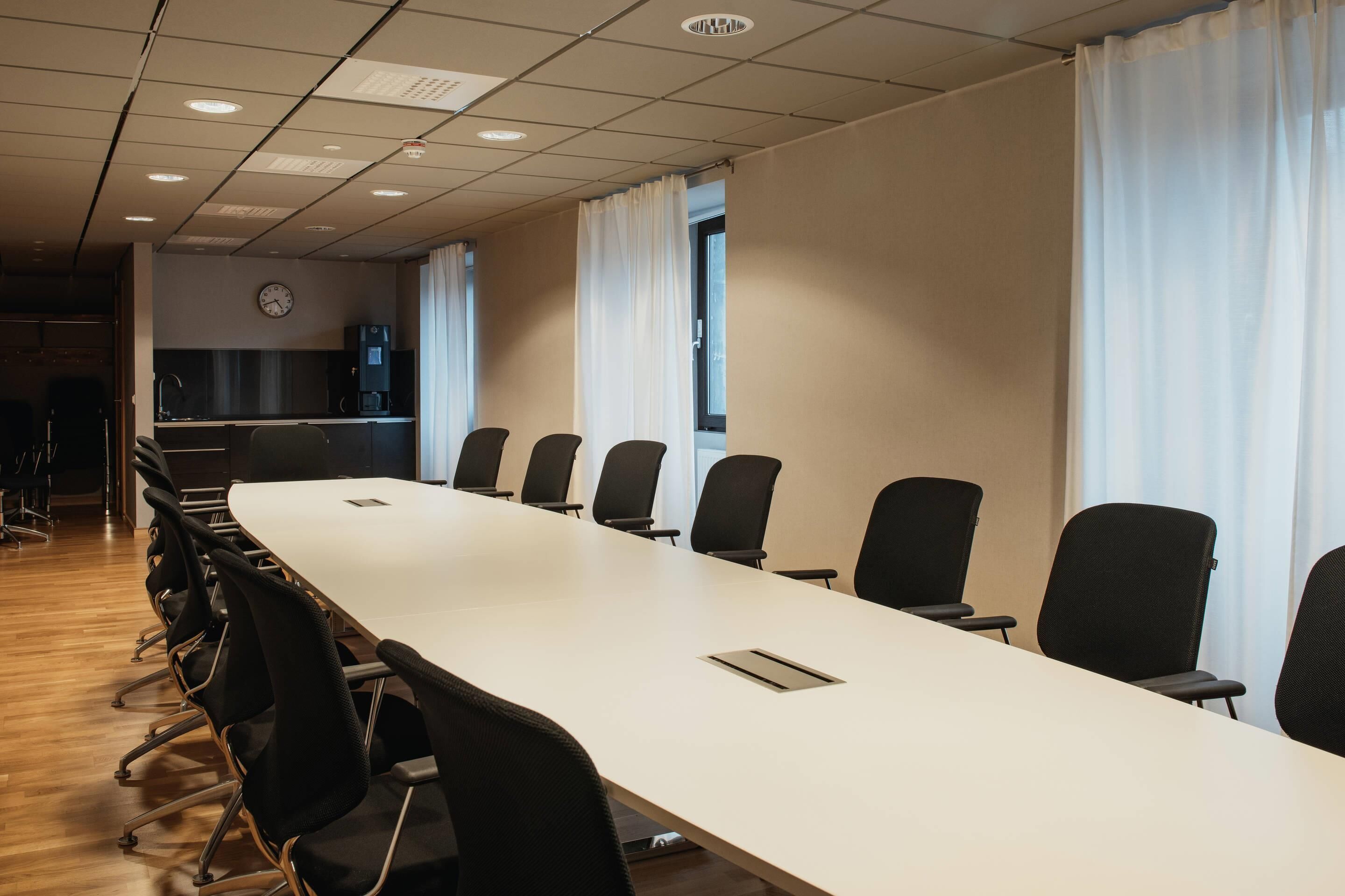 Un salón de conferencias con una larga mesa, sillas negras, un reloj y una zona de cocina al fondo.