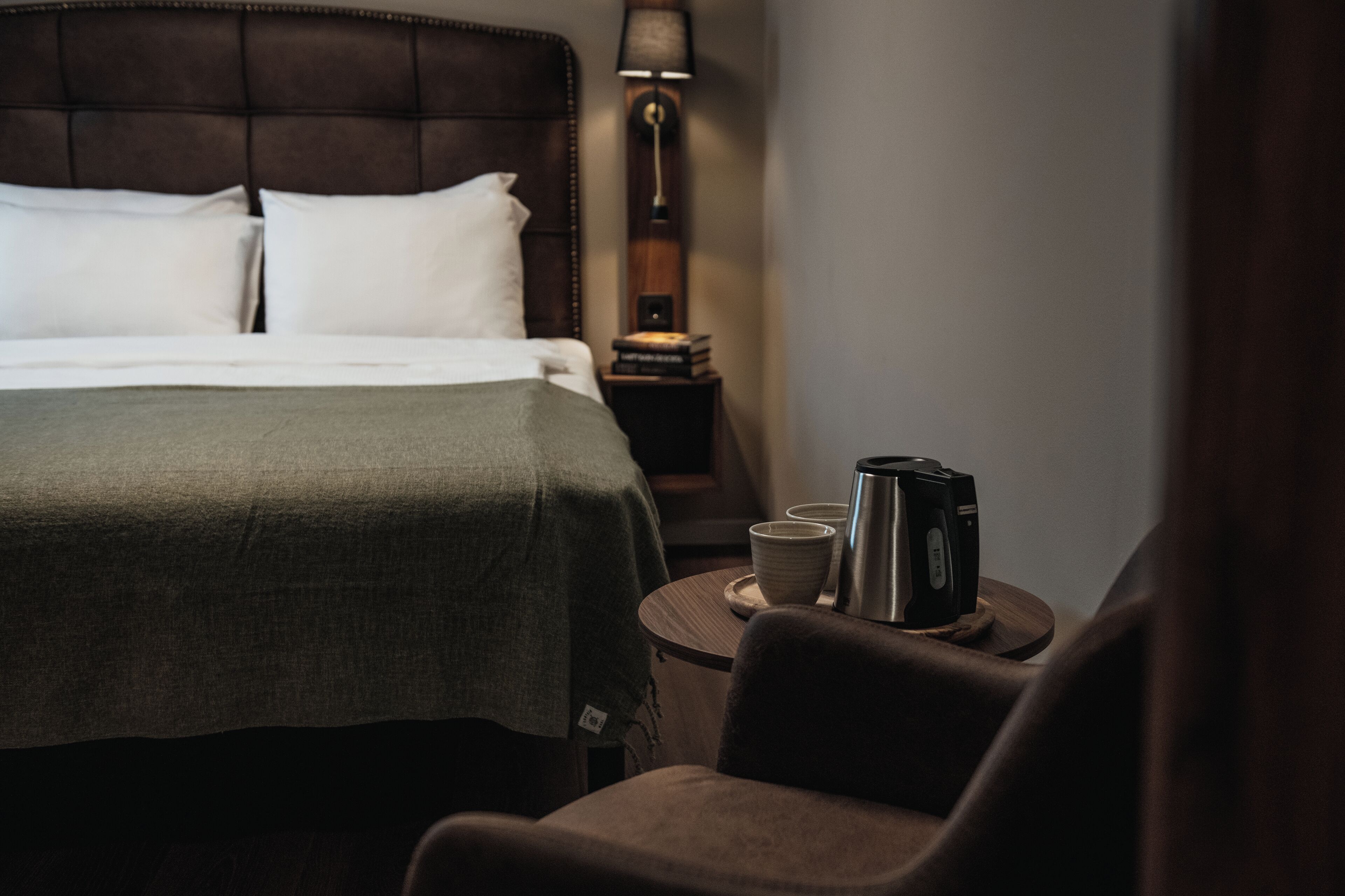 A hotel room with a bed, a bedside table with a lamp and a kettle, and a chair.