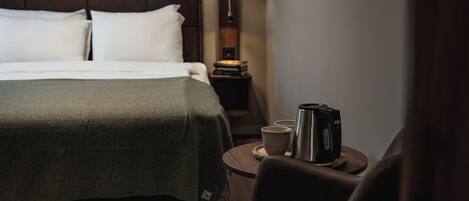 A hotel room with a bed, a bedside table with a lamp and a kettle, and a chair.