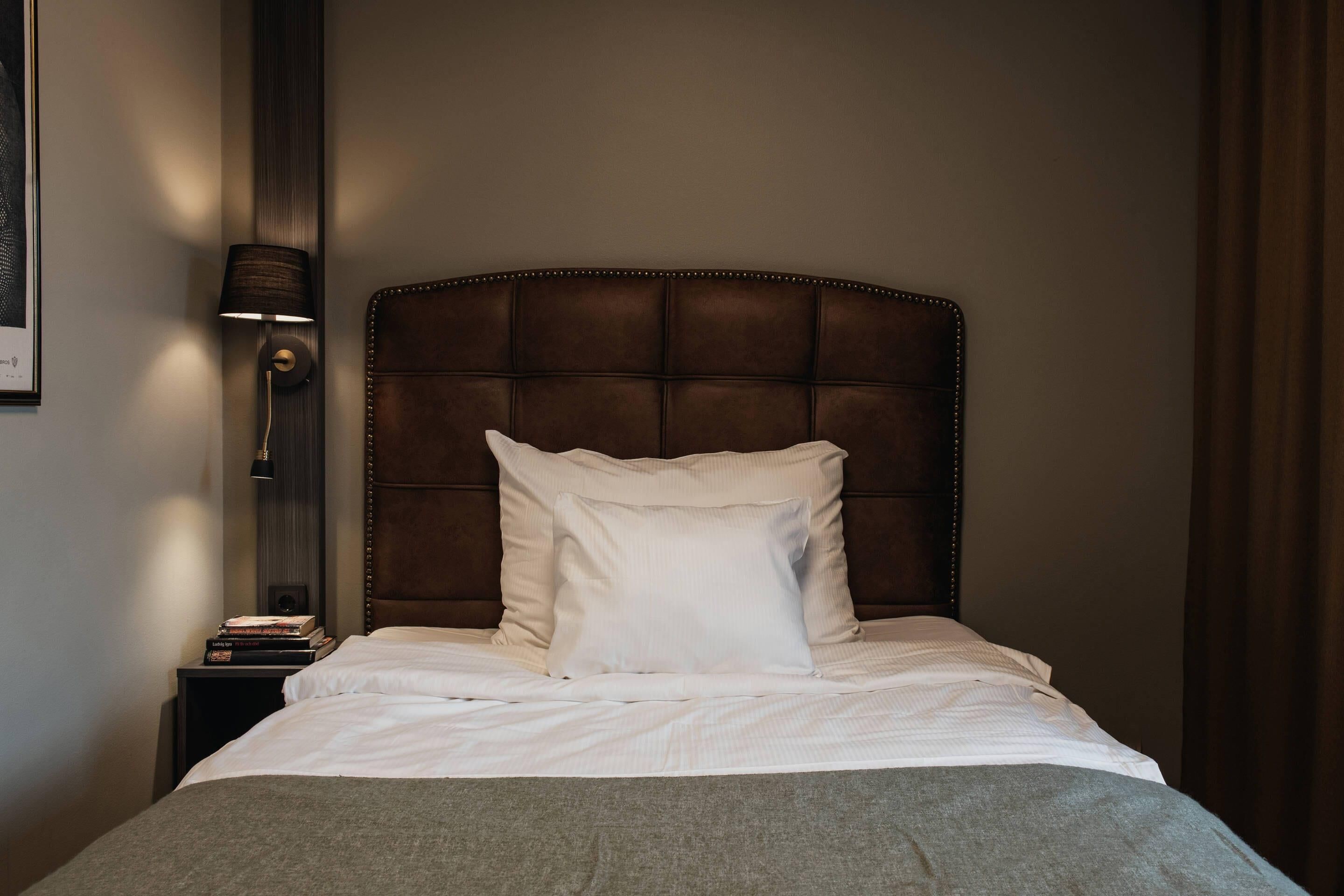 A neatly made bed with a headboard, a bedside lamp, and a stack of books on a nightstand.