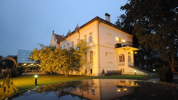 Um edifício grande e histórico com fachada simétrica, entrada central e reflexo em um espelho d'água.