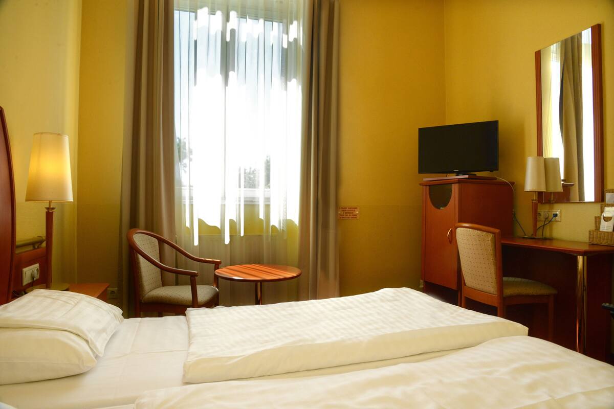 Habitación de hotel con cama, escritorio, silla, televisor y ventana con cortinas.
