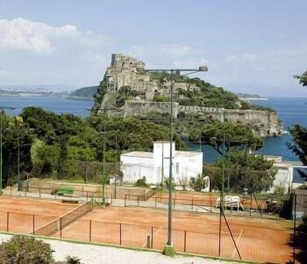 A coastal view with a historic fort on a cliff, a tennis court, and a white building in the foreground. - Parco Cartaromana (Ischia)