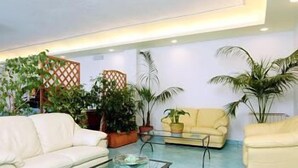 Una sala de estar moderna con un sofá blanco, una mesa de centro de vidrio y un sofá amarillo. Se observan varias plantas en macetas y una estructura de celosía de madera al fondo.