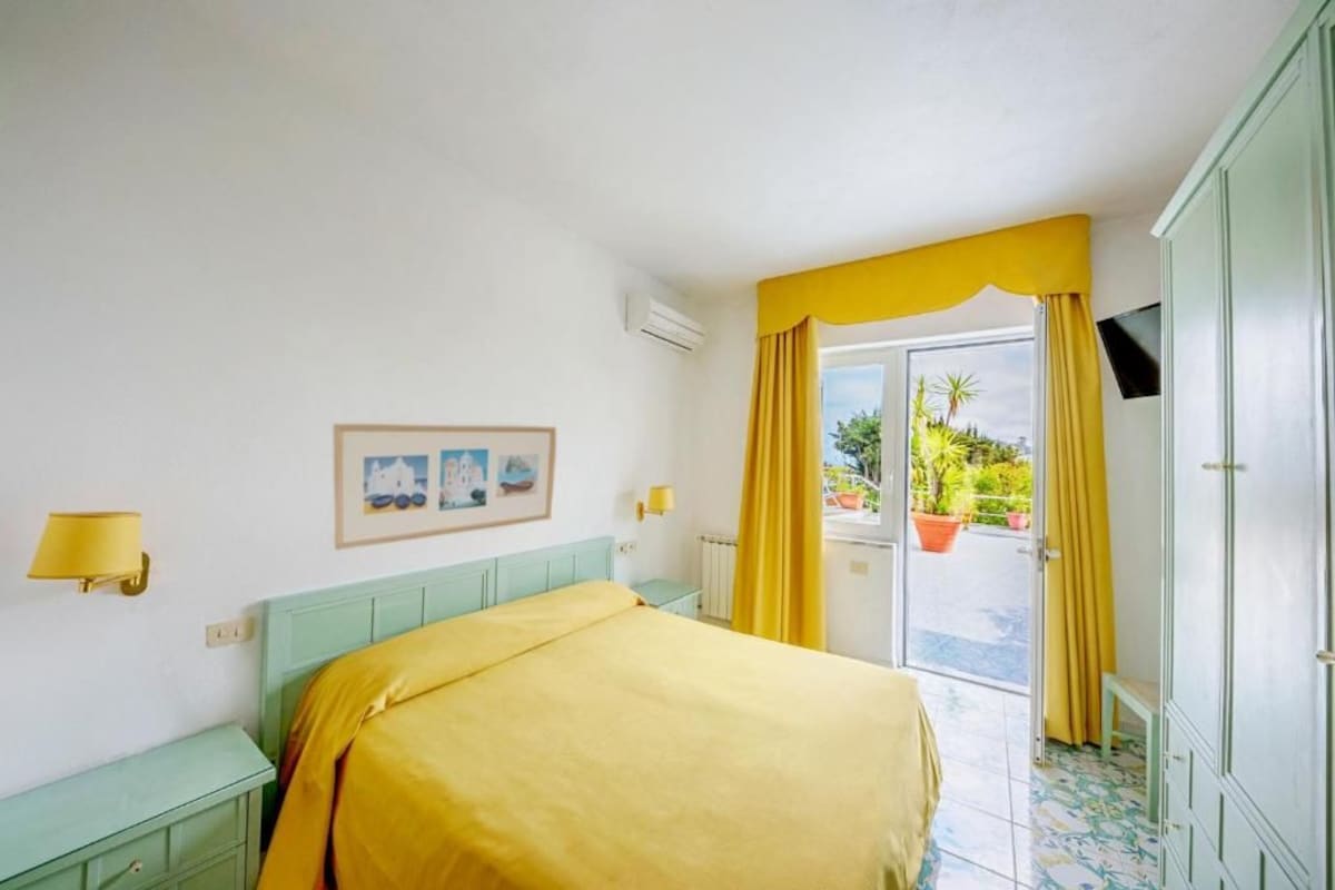 A room with a bed, a desk, a chair, a window with curtains, and a view of the outside.