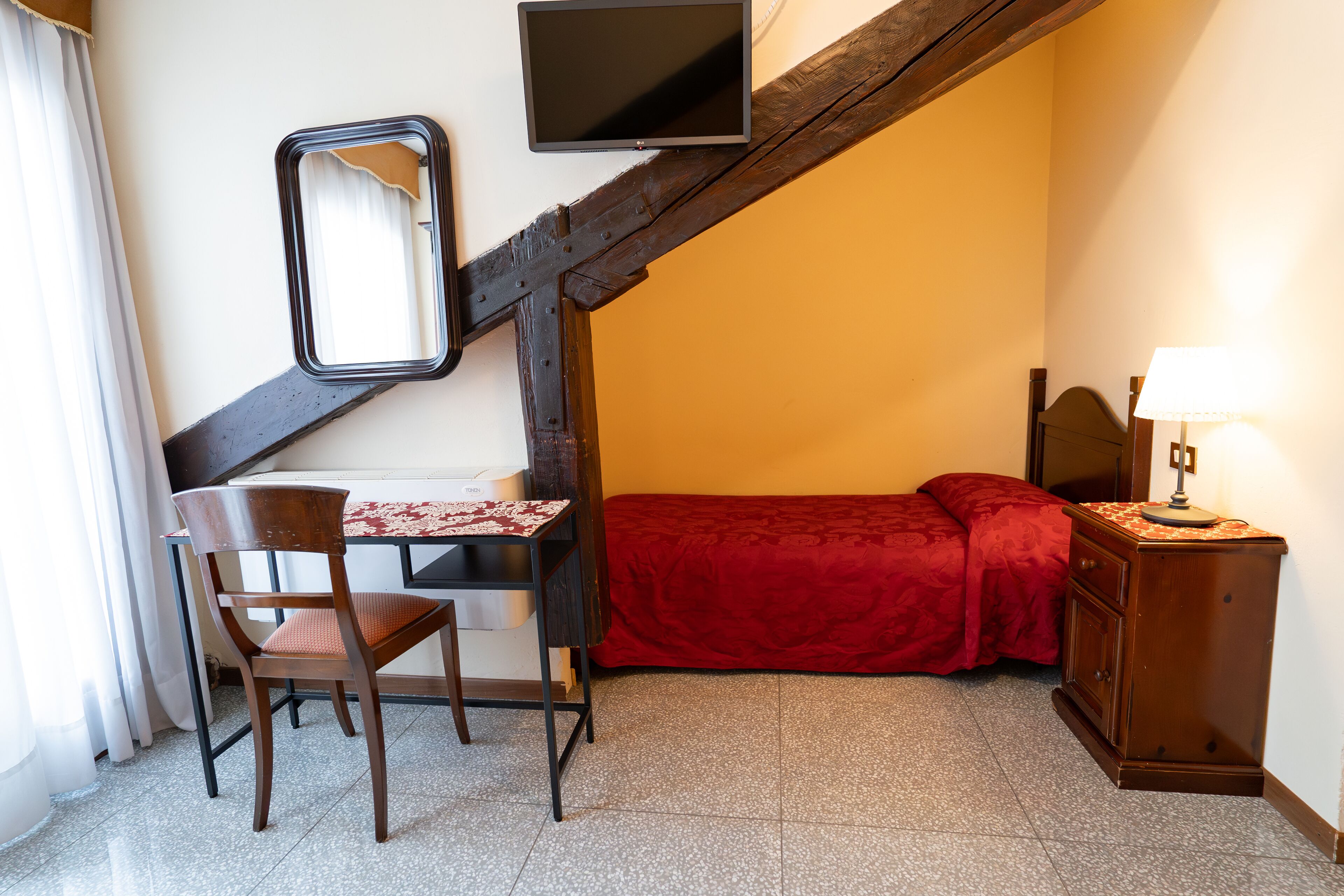 Ein Schlafzimmer mit Bett, Schreibtisch mit Stuhl, an der Wand montiertem Fernseher und Nachttisch mit Lampe.