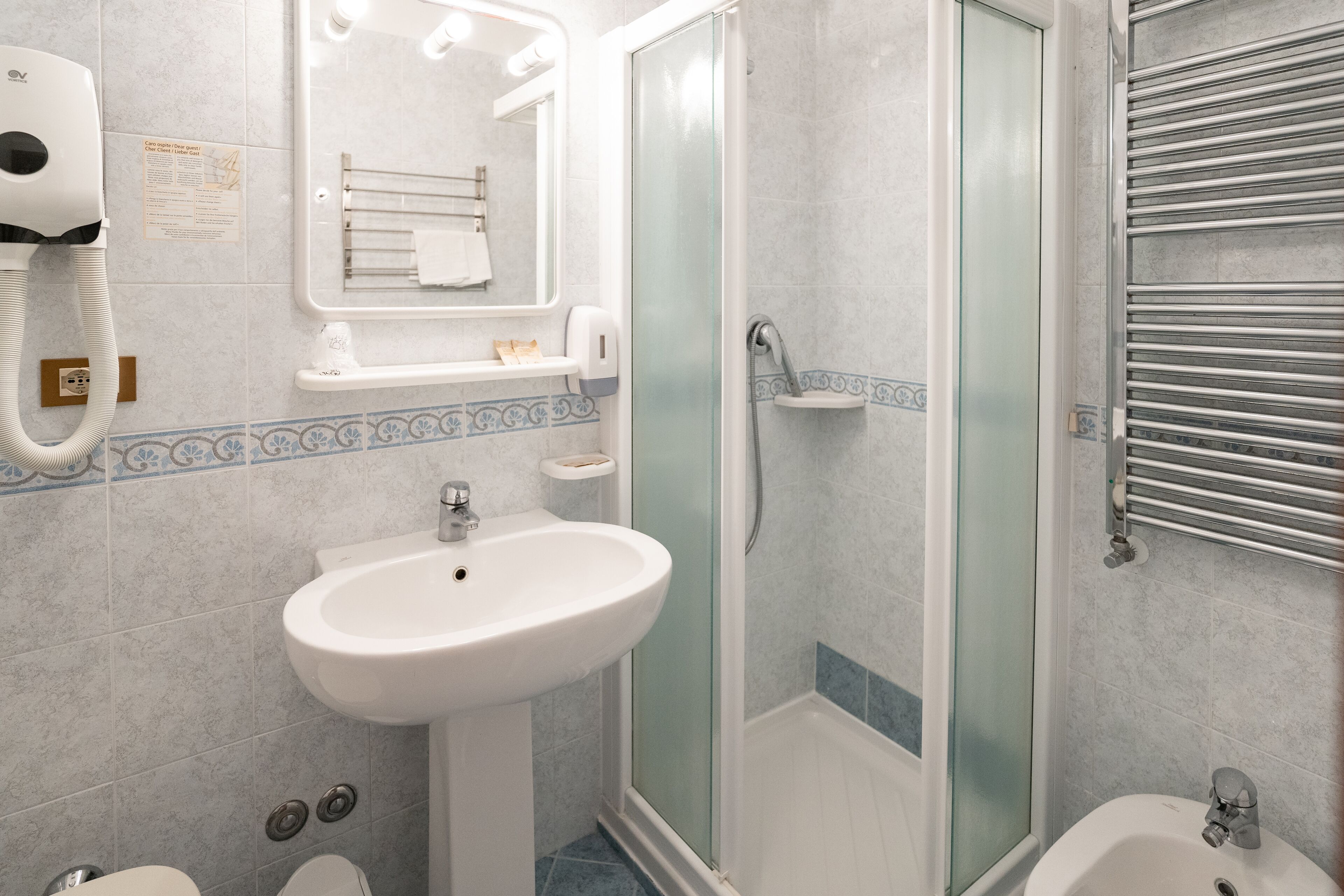 A bathroom with a sink, a glass-enclosed shower, a towel rack, and a hand dryer.