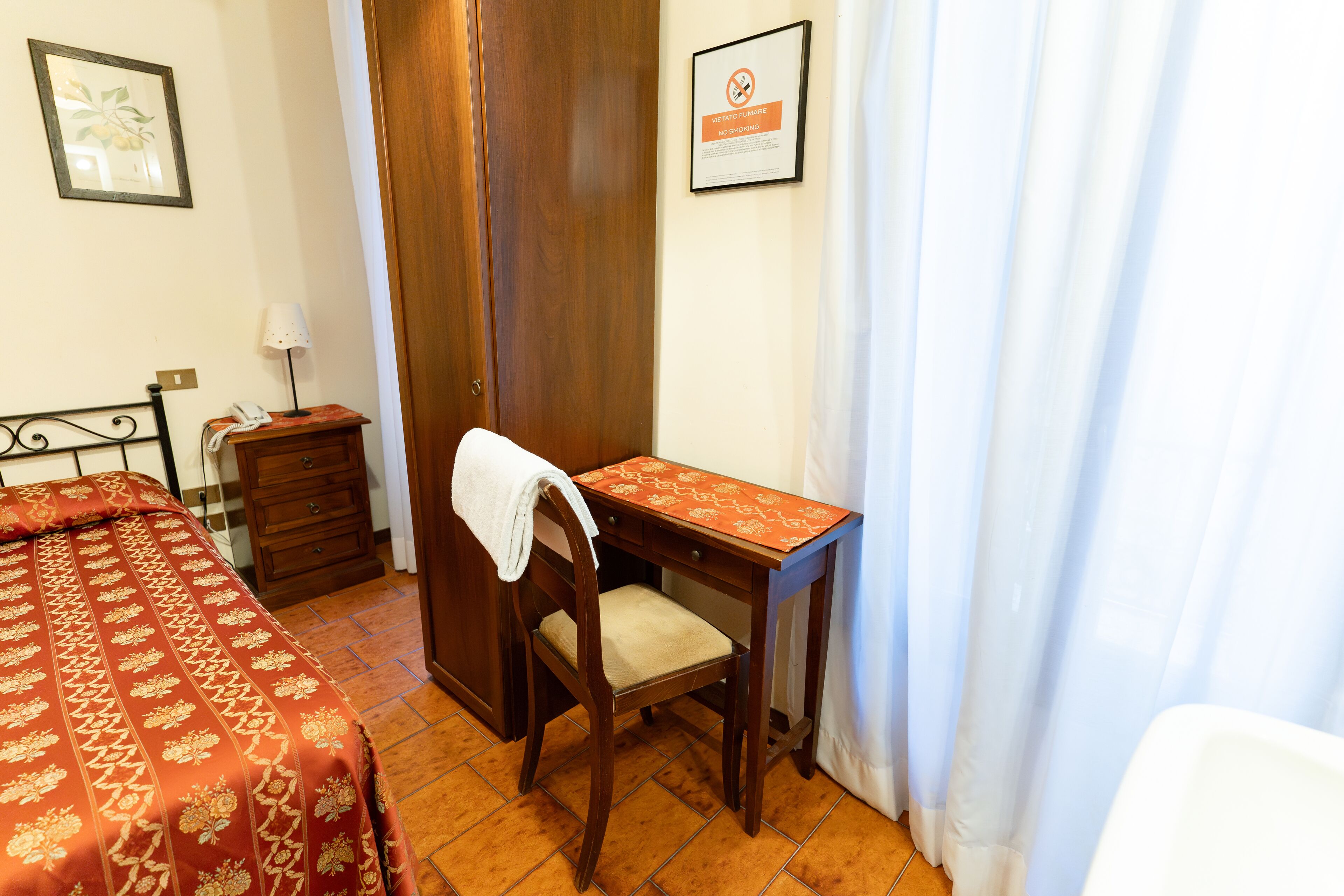 Une chambre d’hôtel avec un bureau en bois, une chaise sur laquelle est posée une serviette, une table de chevet avec une lampe, un lit recouvert d’une courtepointe rouge et un panneau encadré accroché au mur.