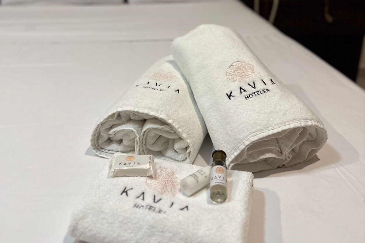 A neatly made bed with white linens, a bottle of lotion, and a folded towel with 'KAVIA HOTEL' branding.
