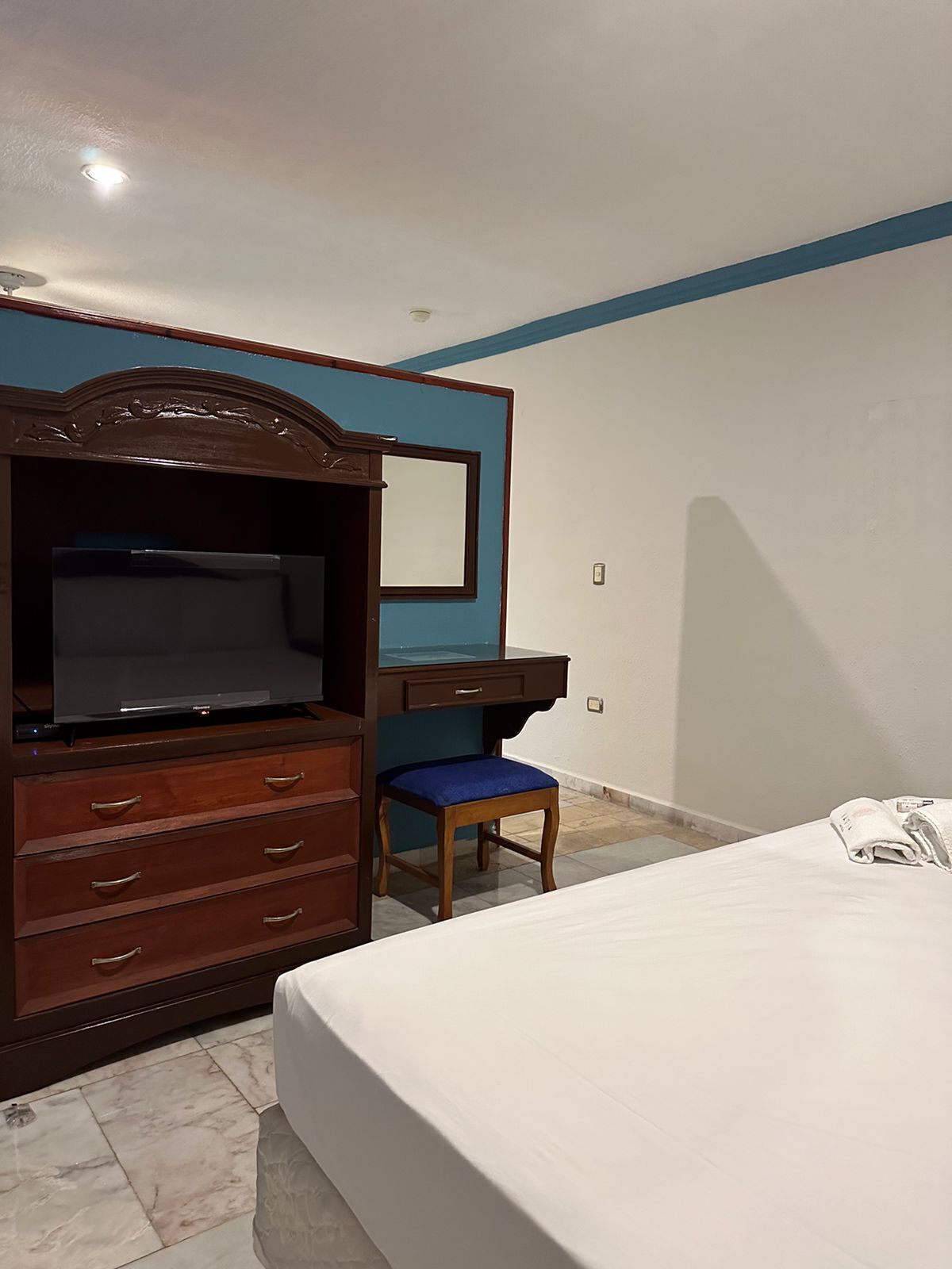 A bedroom with a bed, a wooden dresser with a television, a desk, and a chair.