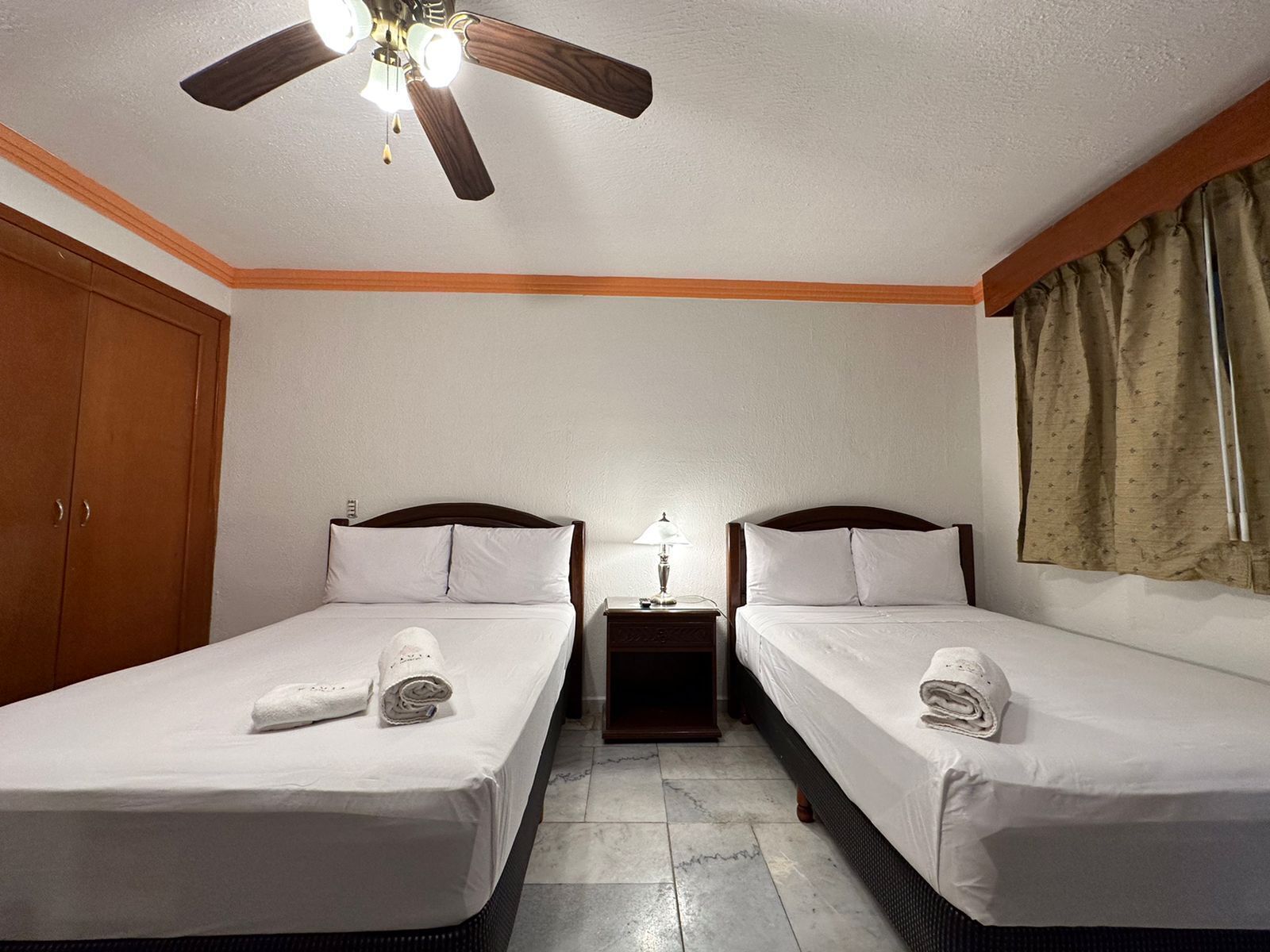 A hotel room with two single beds, a wooden wardrobe, a ceiling fan, a nightstand with a lamp, and a window with curtains.