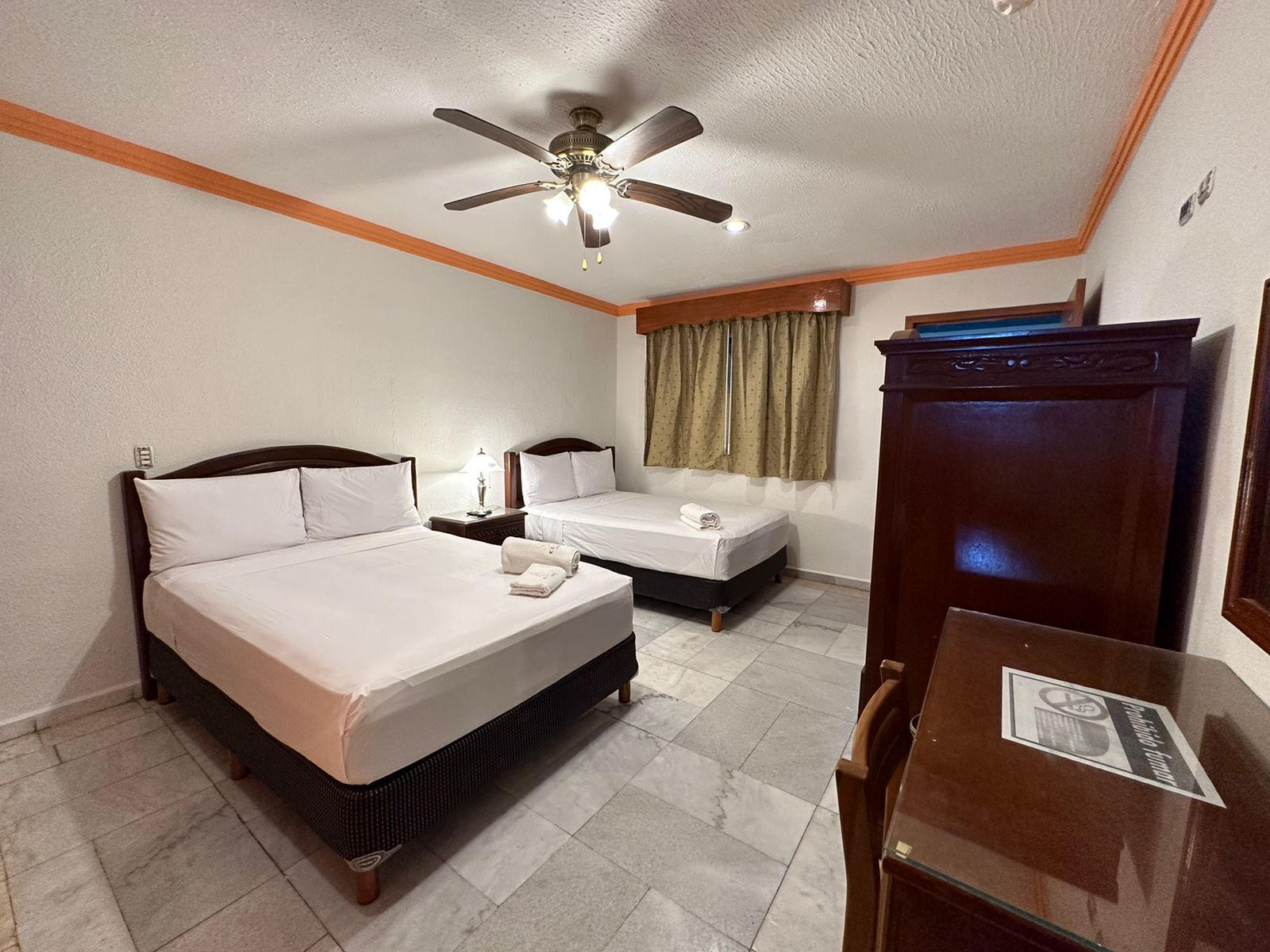 A hotel room with a large bed, a smaller bed, a desk, a wardrobe, a ceiling fan, and a window with curtains.