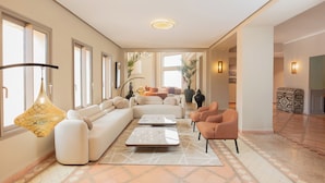 A spacious living area with a large sofa, two coffee tables, and a patterned rug. There are multiple windows providing natural light, and a decorative hanging light fixture. - Kenzi Azghor (Ouarzazate)