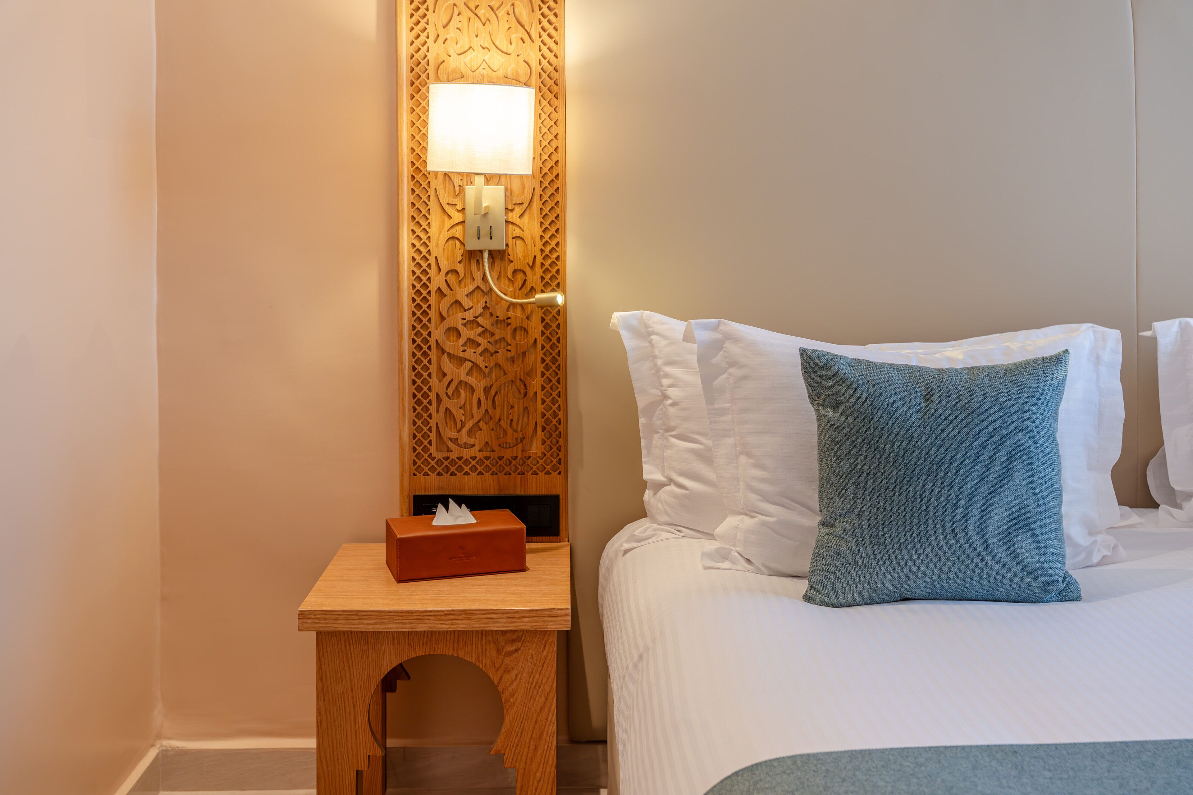 A neatly made bed with white and blue pillows, a bedside table with a tissue box, and a wall-mounted lamp.