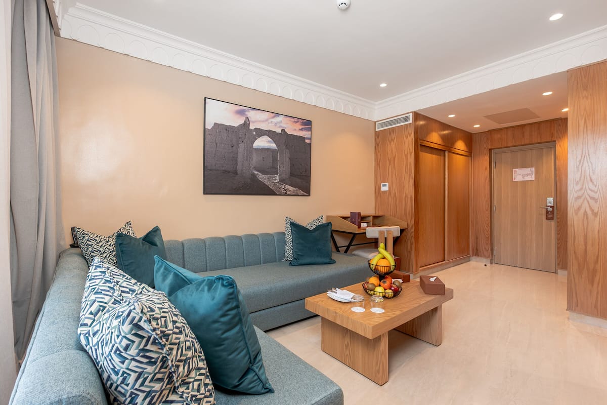 A modern hotel lobby with a grey sofa, teal pillows, a wooden coffee table with fruit, and a large wall picture.