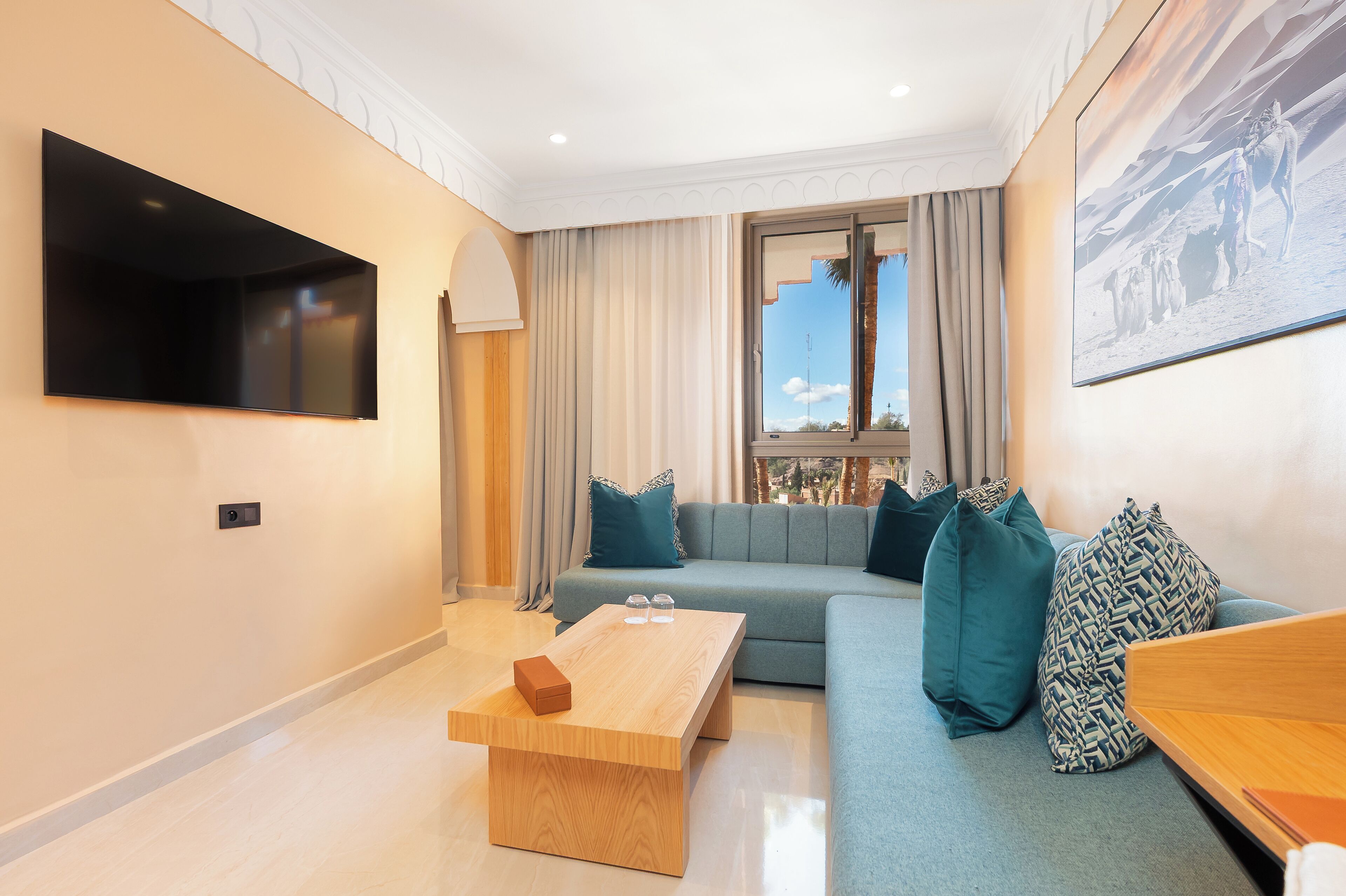 Ein modernes Wohnzimmer mit einem Flachbildfernseher, einem Holztisch, einem grauen Sofa mit gemusterten Kissen und einem großen Fenster mit Vorhängen.
