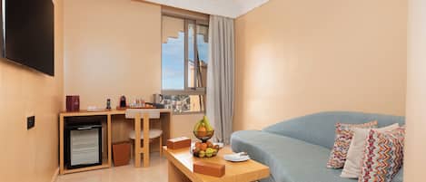 A compact living space with a sofa, a small table with fruits, a mini-fridge, a desk, and a TV mounted on the wall.