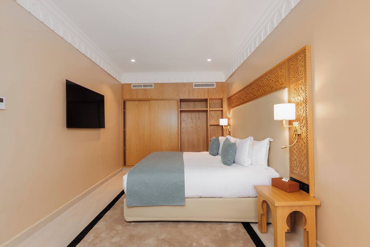 A hotel room with a large bed, wooden headboard, bedside lamps, and a flat-screen TV mounted on the wall.