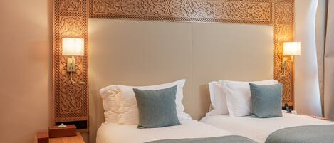 A hotel room with two beds, each with white linens and blue pillows. There are wall-mounted lamps on either side of the beds.