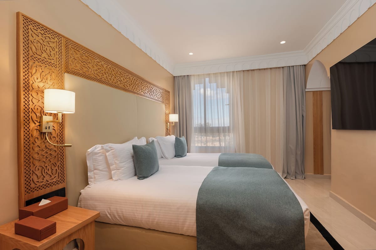 A hotel room with two beds, a wooden headboard, a bedside table with a lamp, and a large window with curtains.
