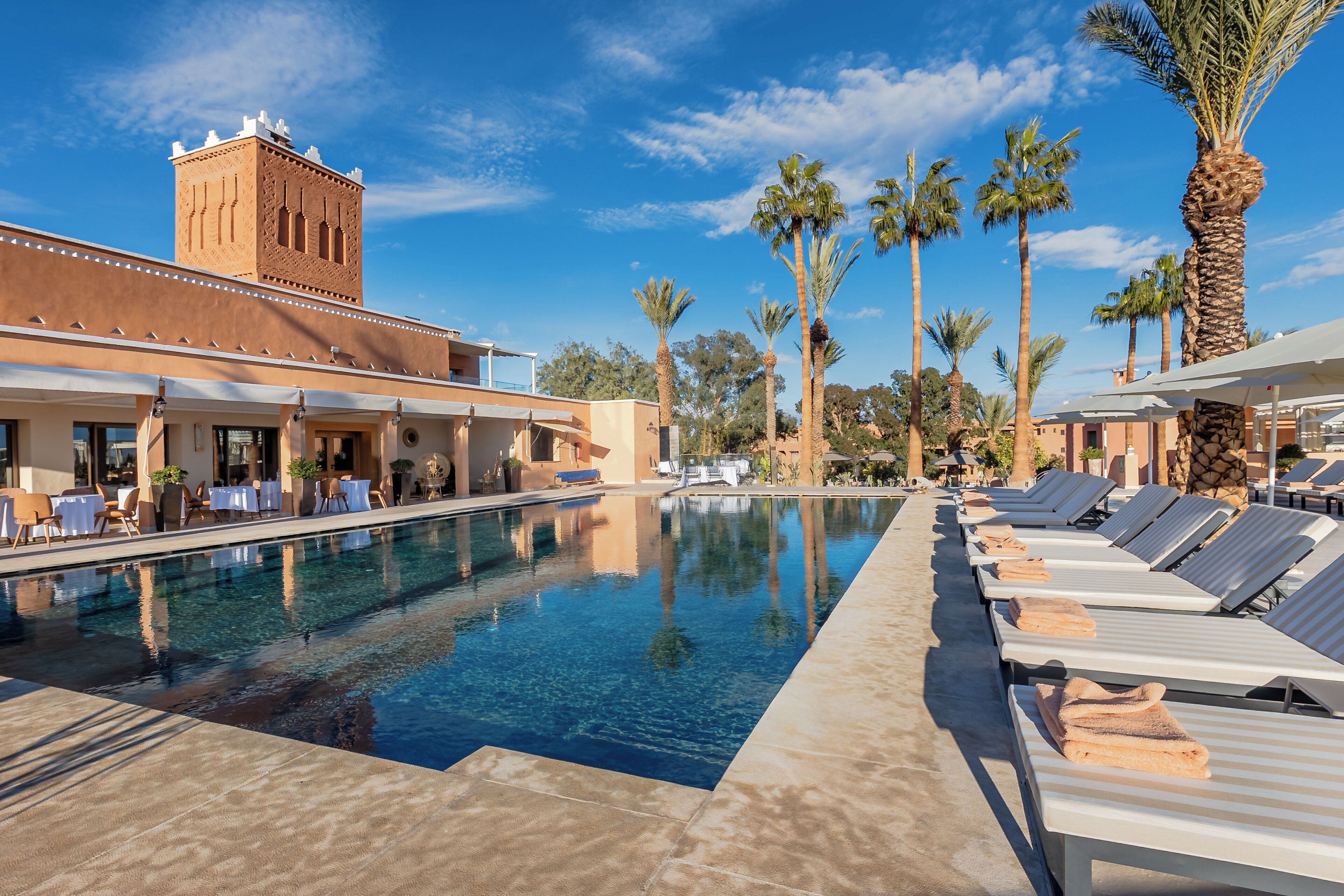Ein Pool mit Liegestühlen und Palmen, ein Gebäude mit einem Turm, und ein klarer, blauer Himmel.