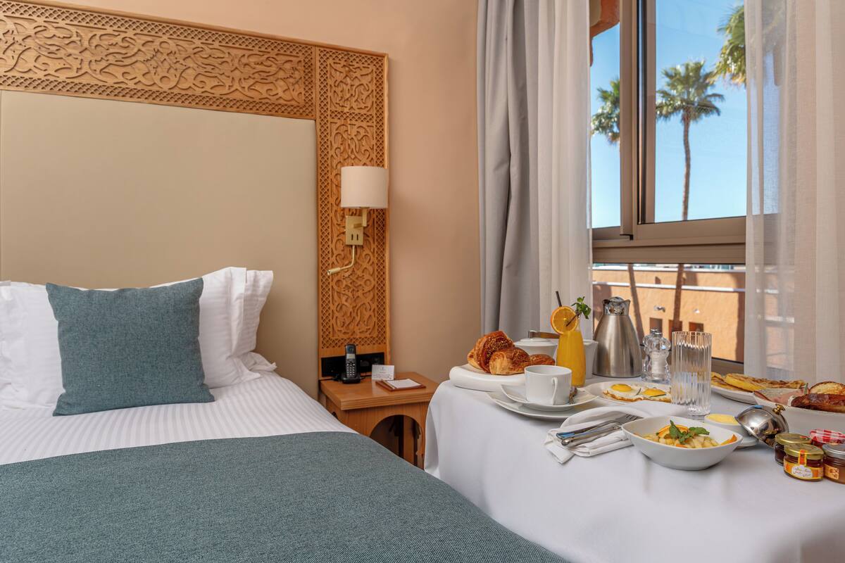 A hotel room with a bed, a dining table with breakfast items, a window with a view of palm trees, and a decorative headboard.